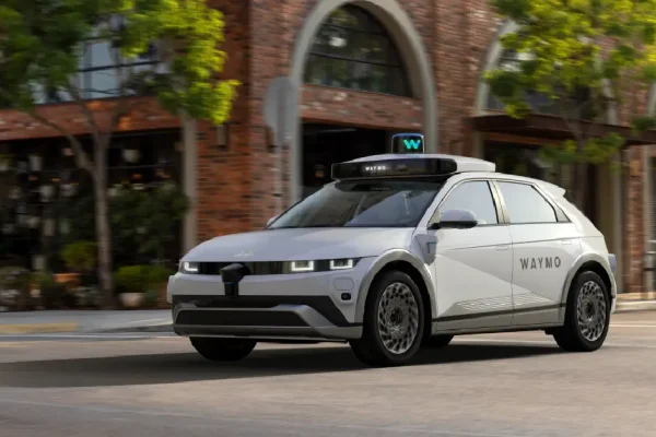 Waymo self-driving car driving on a city street in front of brick buildings and trees
