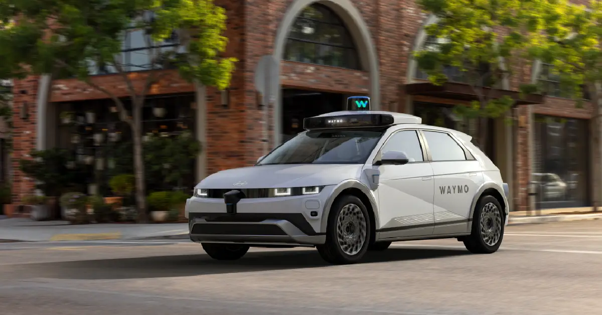 Waymo self-driving car driving on a city street in front of brick buildings and trees