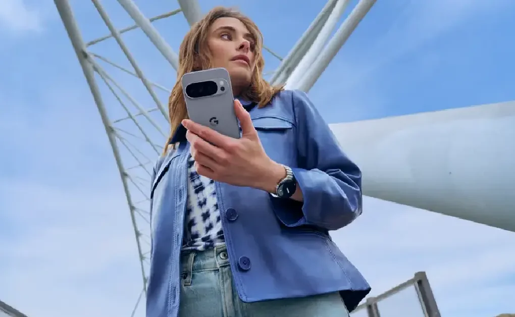A woman in a blue jacket holds a Google Pixel phone and wears a Pixel Watch outdoors beneath a modern white structure against a blue sky
